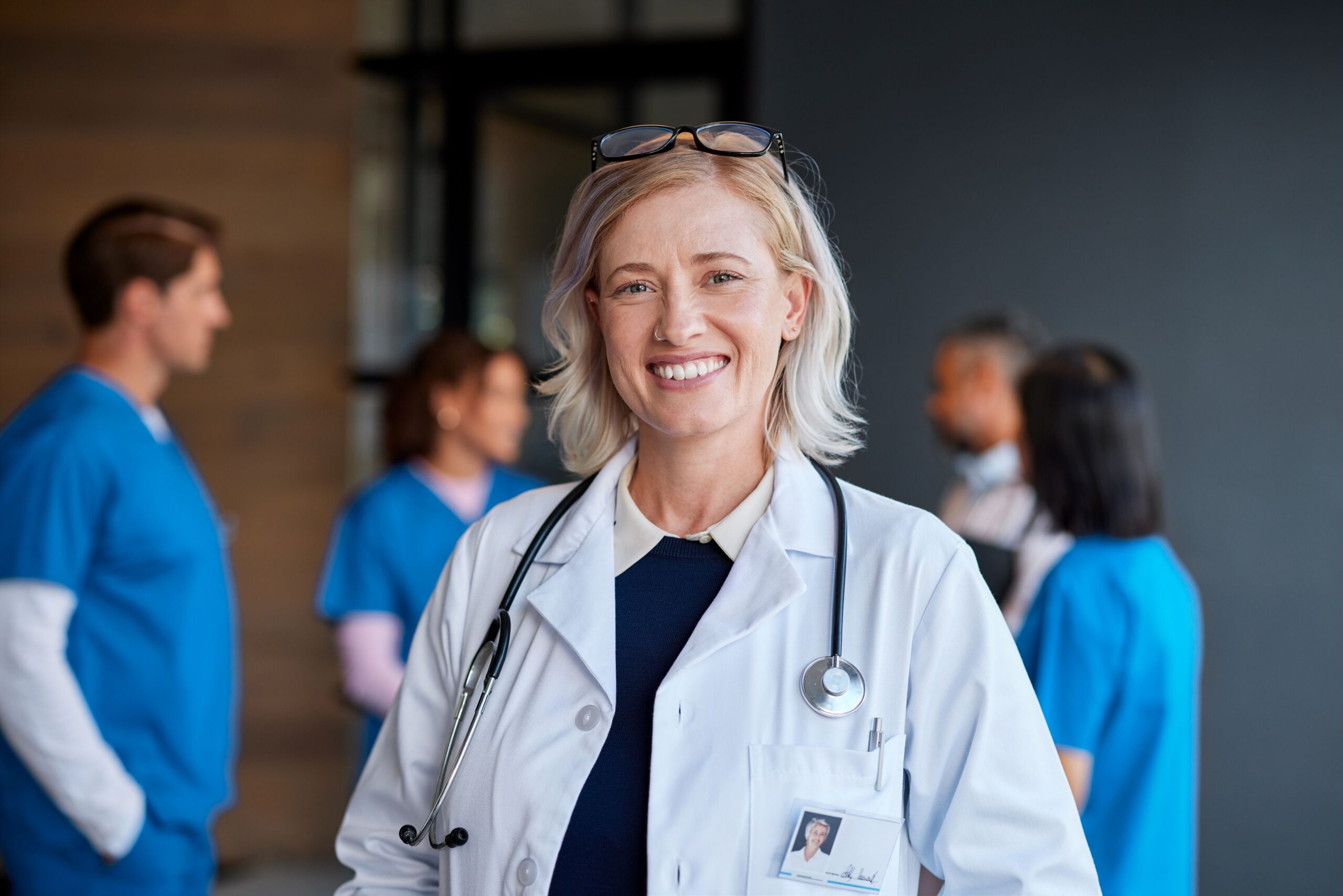 Best wordpress plugins, image of a smiling doctor with short blonde hair wearing a white doctor’s coat, stethoscope around her neck, a blue undershirt and glasses on her head with a group of medical practitioners in the background.