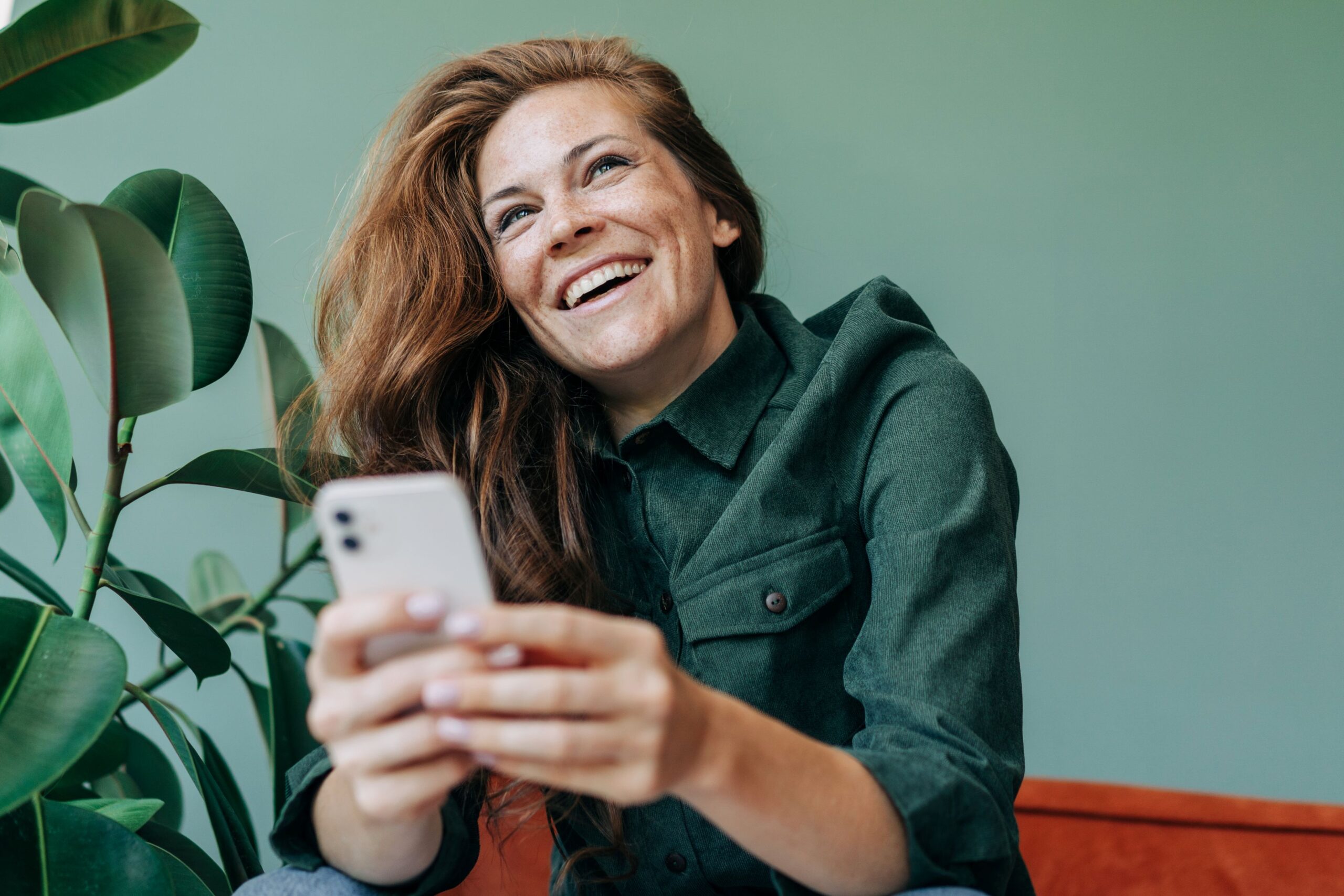 Best wordpress plugins, image of a smiling woman with long red hair wearing a green buttoned-down shirt sits on an orange couch holding her smartphone with both hands.