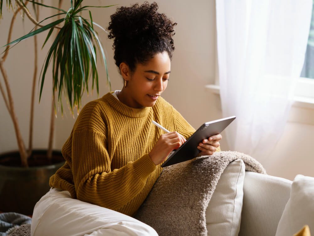 Ebook in libraries, image of a woman wearing a mustard yellow sweater and white pants sits on a white couch while holding a digital tablet in her left hand and a stylus pencil in her right hand.