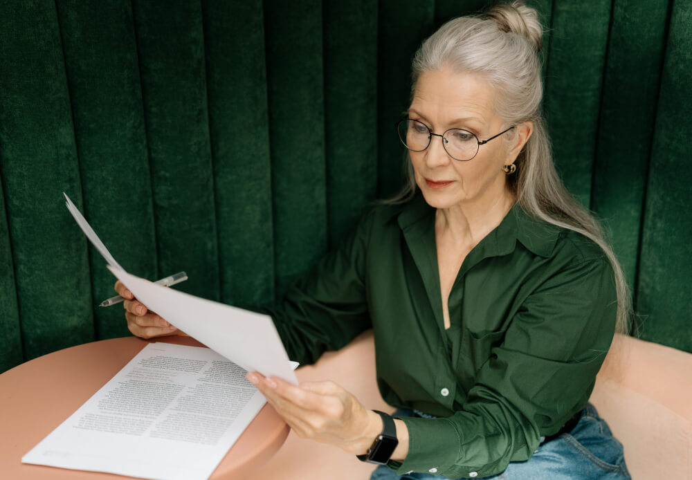 Ebook in libraries, image of a woman with long gray hair, wearing a dark green blouse and jeans, examines large sheets of paper with print on them.