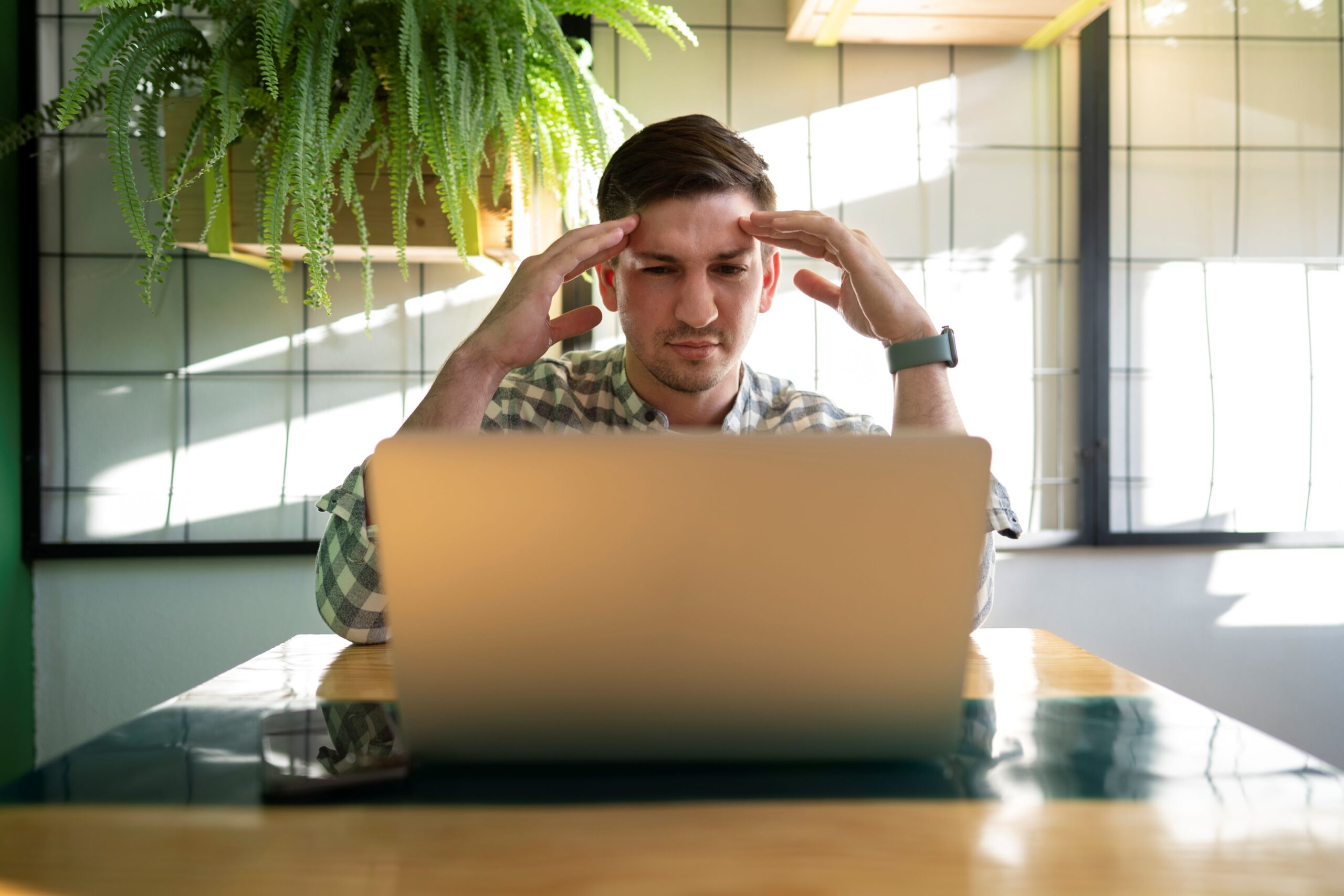 AI Spambots, image of a frustrated man with his hands touching his forehead, sitting at a table with his laptop in front of him.