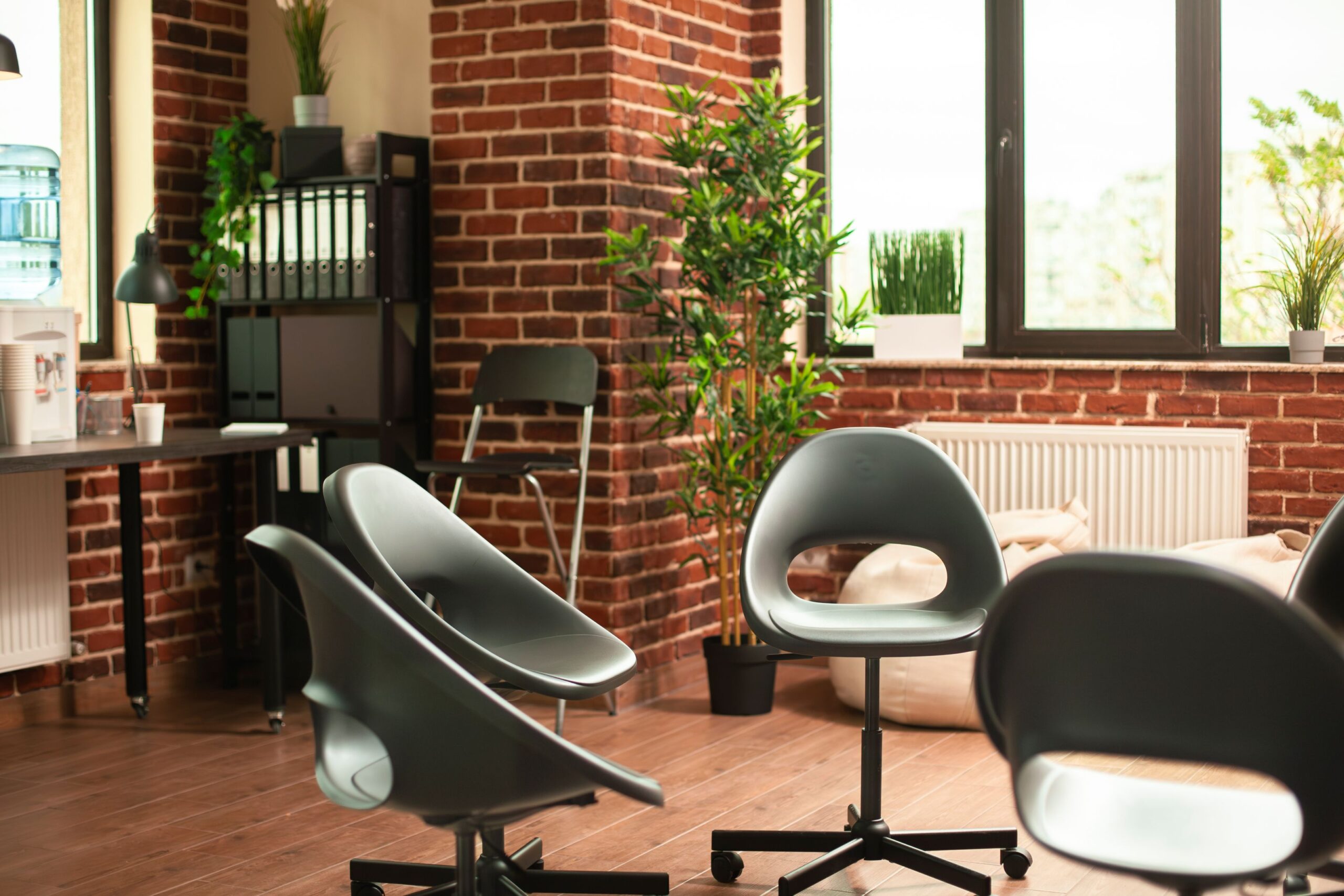 AI Spambots, image of a therapy office with brick walls, large windows, a desk and a set up of 5 green rolling chairs set up in a circle.