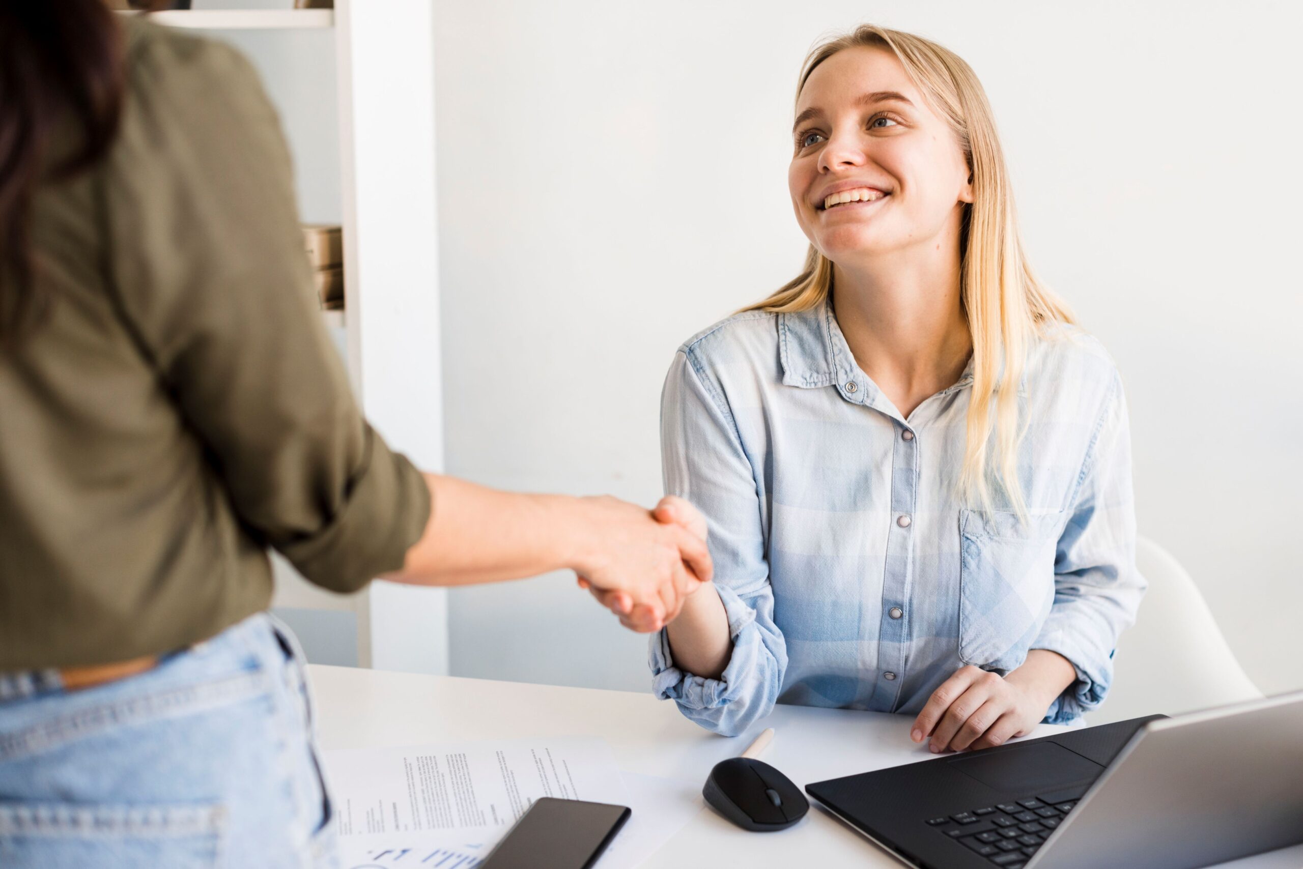 AI Spambots, image of a young woman sitting at a white desk with a laptop in front of her shaking hands with a woman standing on the other side of the desk.