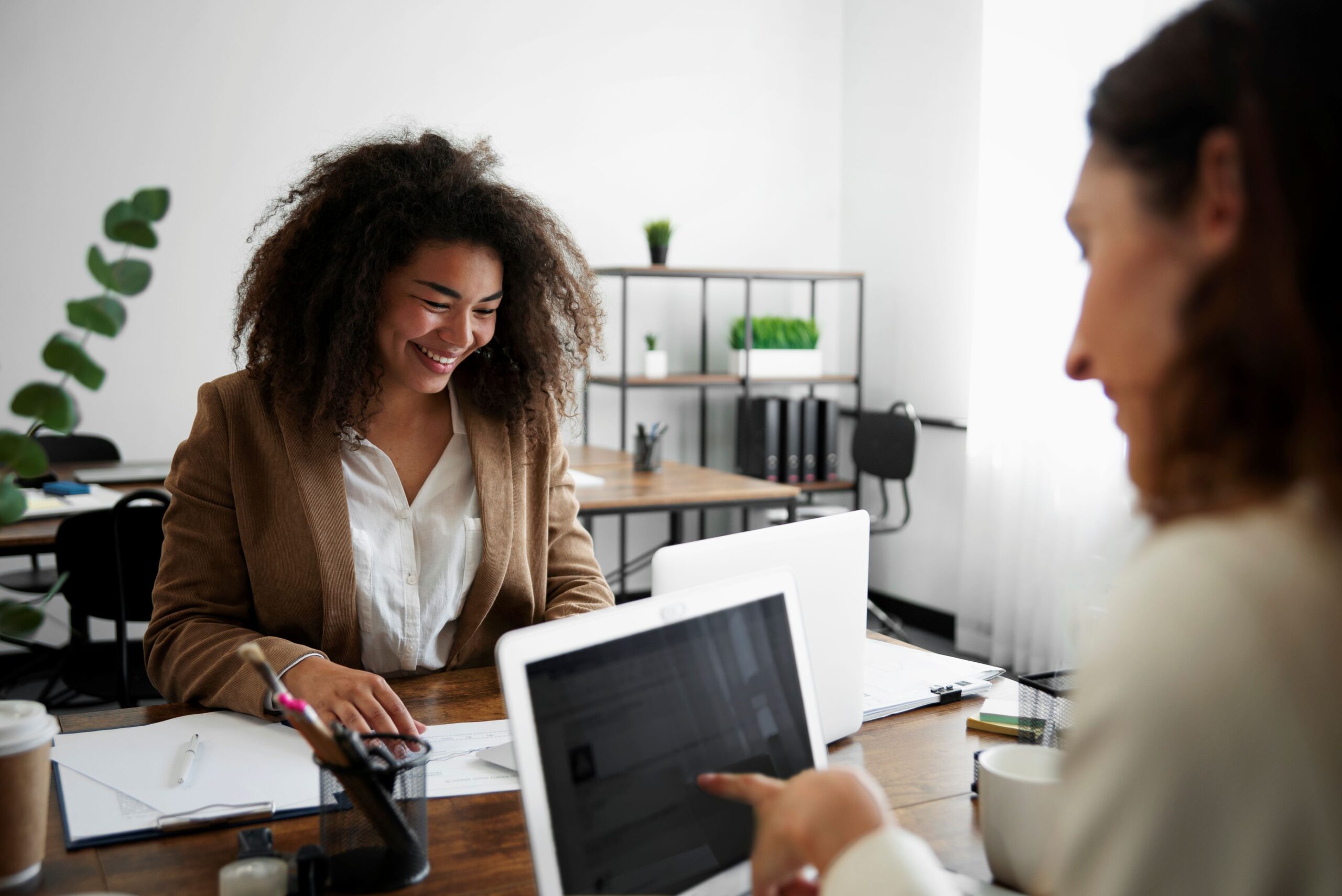 AI Spambots, image of two professional women sitting across from each other at a wooden table filled with two laptops and office supplies.