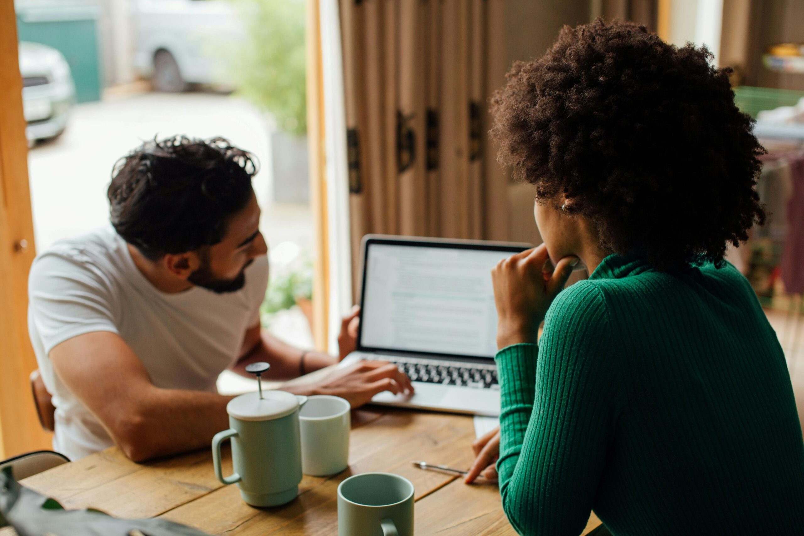 Defend Your WordPress, image of a woman wearing a green sweater and a man wearing a white t-shirt sitting across from each other at a wooden table filled with a coffee press and two coffee cups, both looking at an open laptop.