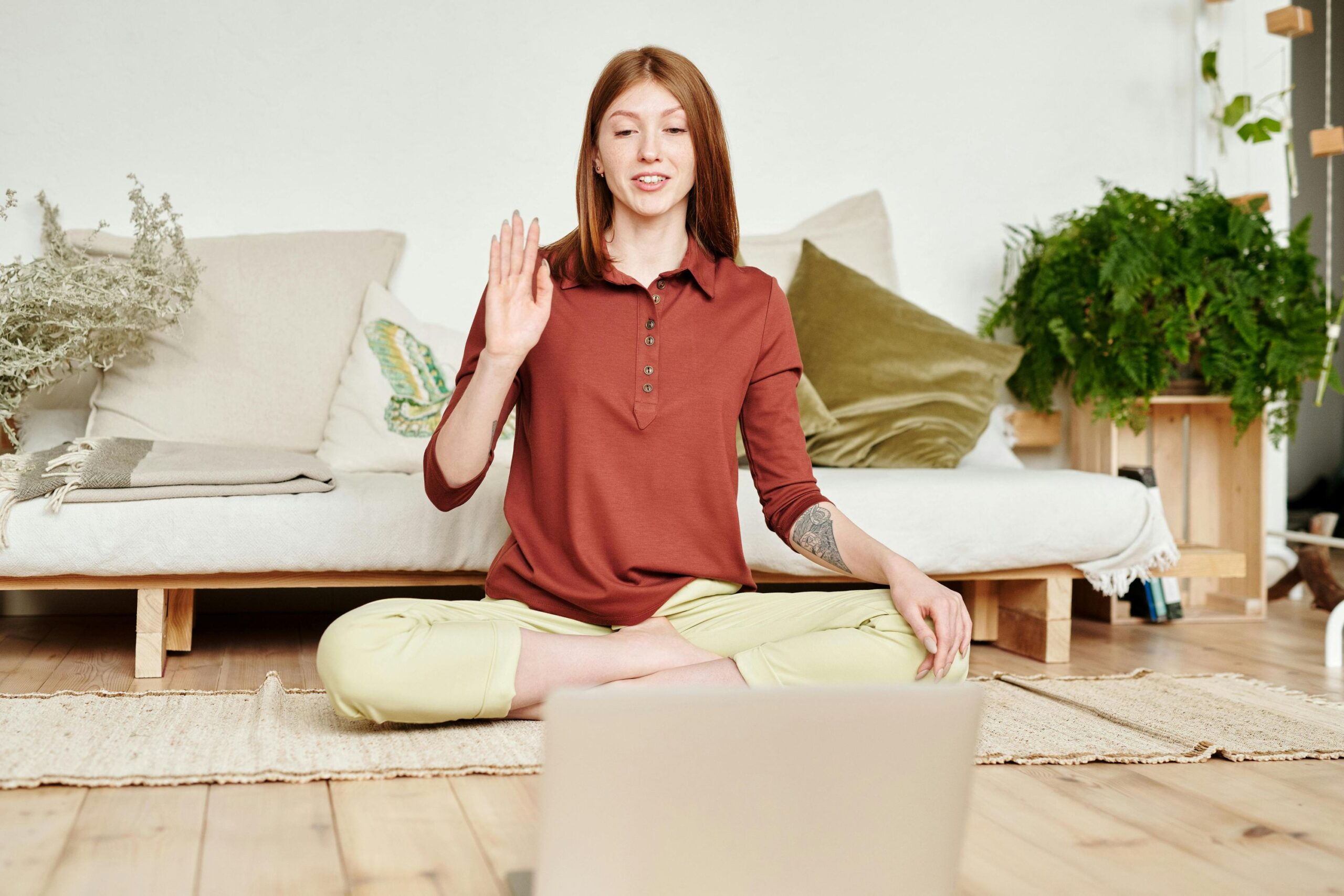 Defend Your WordPress, image of a woman wearing a collared, button-down red blouse and yellow pants sitting on a mat on the floor across from an open laptop, waving hello at the laptop.