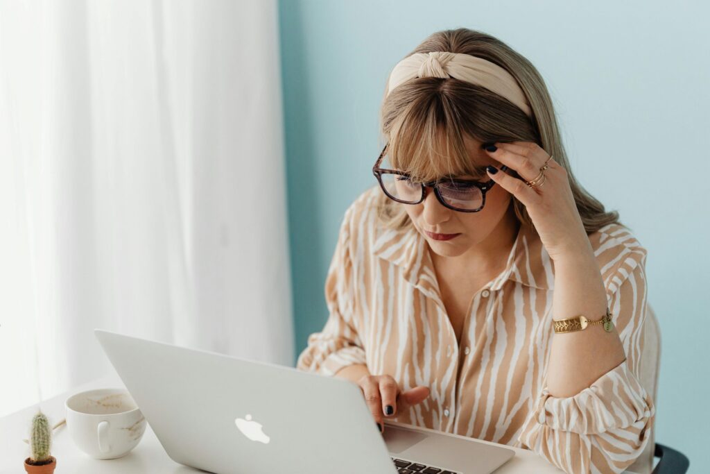 fake cloudflare malware, image of a woman sitting at a table in front of a laptop with an empty coffee mug to the side and her left hand touching her forehead.