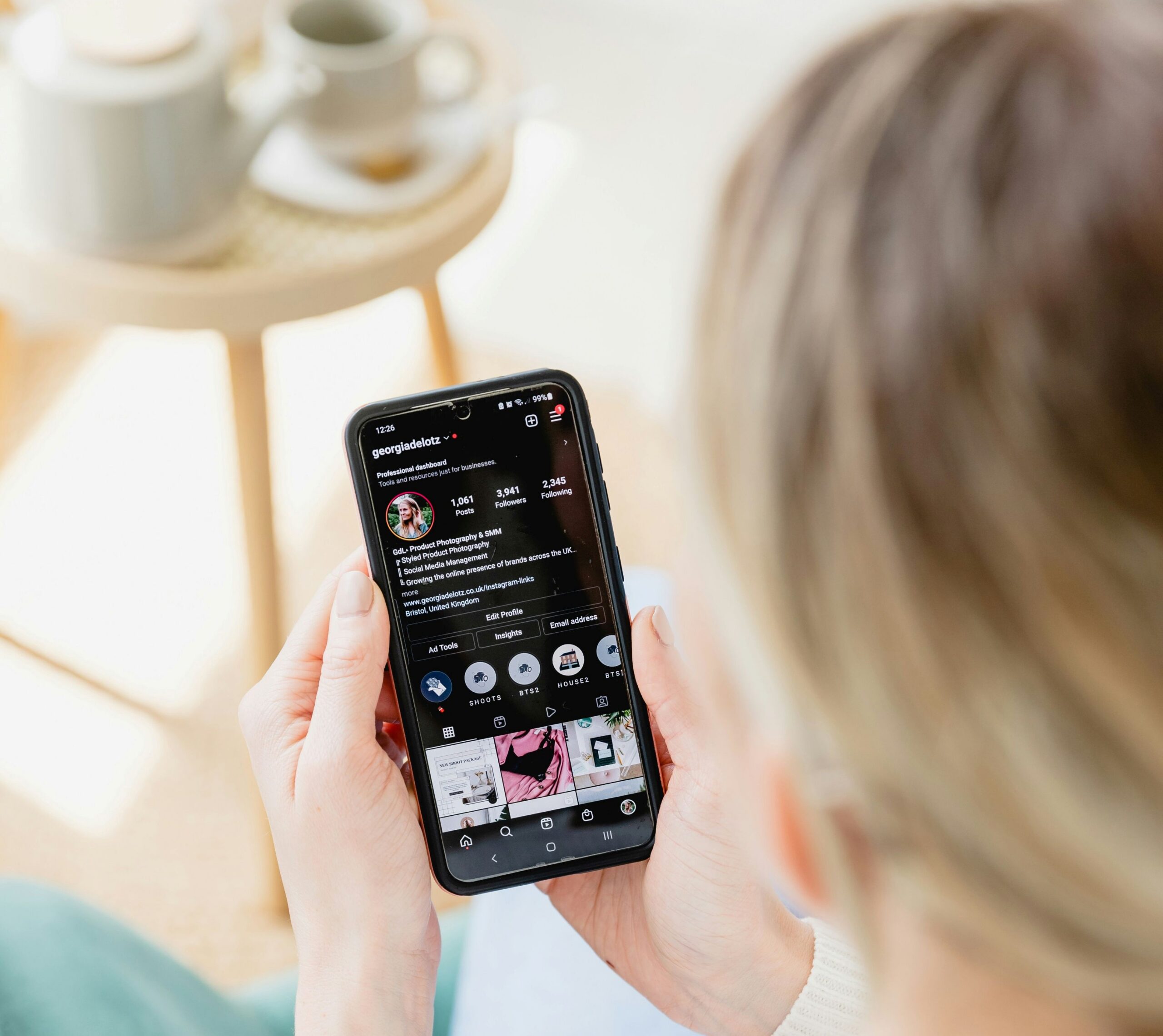 a person in a cream sweater scrolling through an Instagram business profile on their phone, with a light-filled room in the background. Linktree Alternatives.