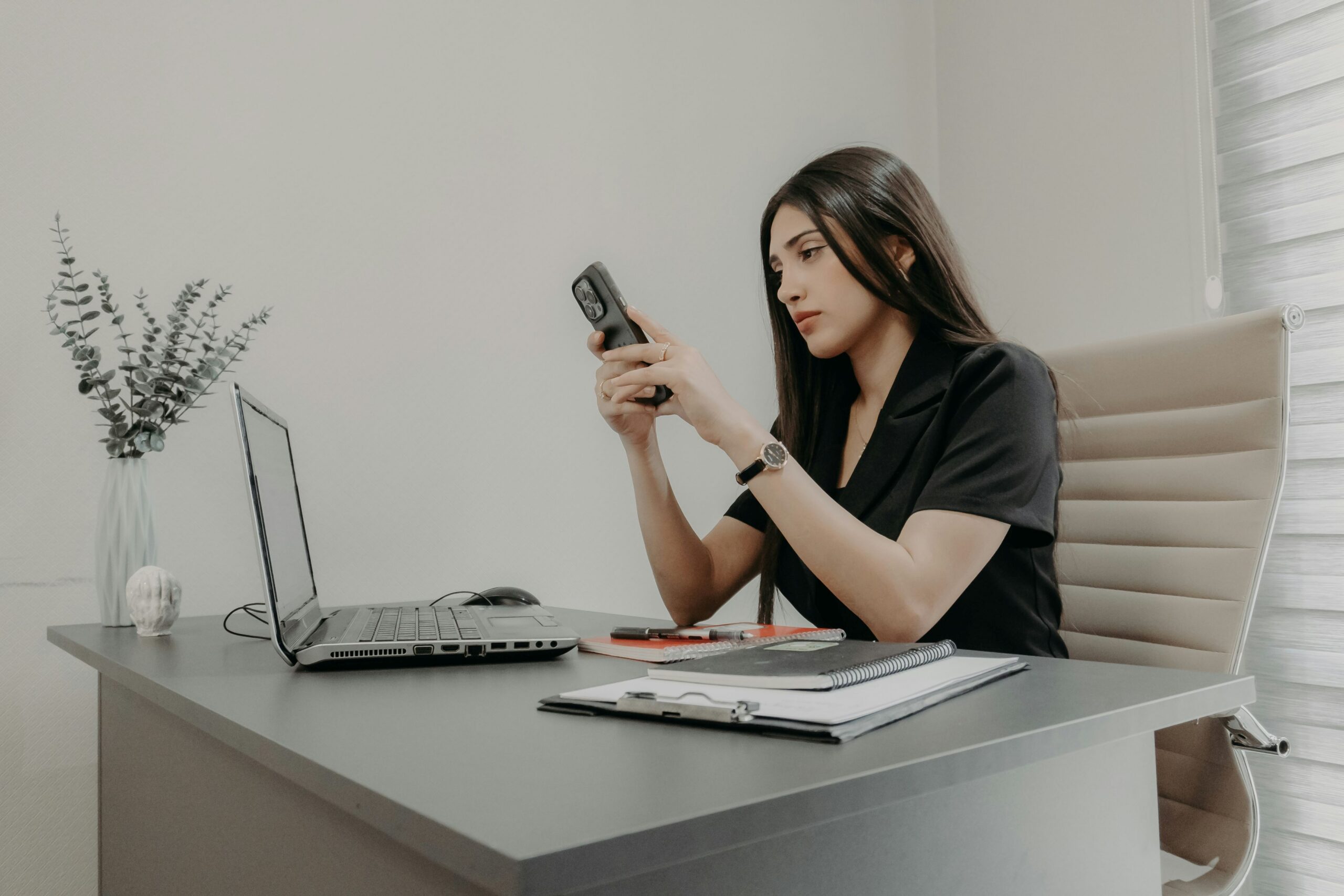 A woman in a black blazer sits at a desk reviewing her phone, with a laptop, notebook, and small eucalyptus plant nearby. Linktree Alternatives.
