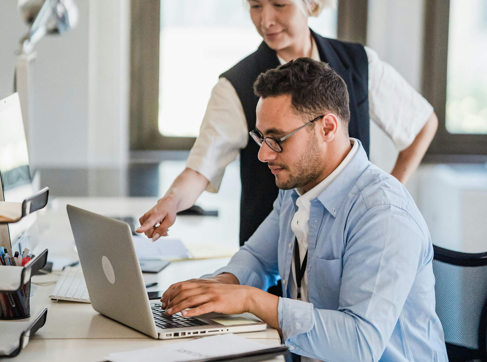 Image of a man sitting at a laptop with a woman standing behind him and pointing at the screen.