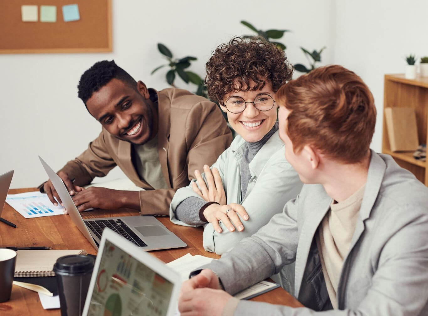 Image of a marketing team of three young professionals sitting and talking at a wood table with laptops.