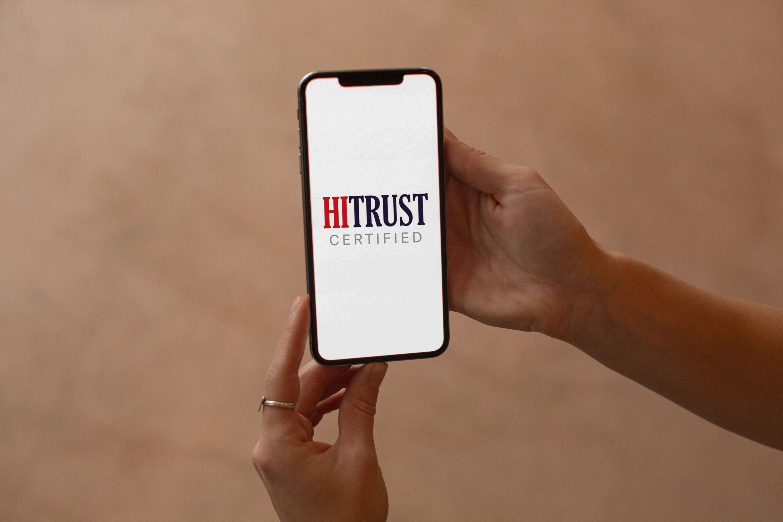 A woman holds her cellphone with both hands, on the screen is a HiTrust certification for security controls.
