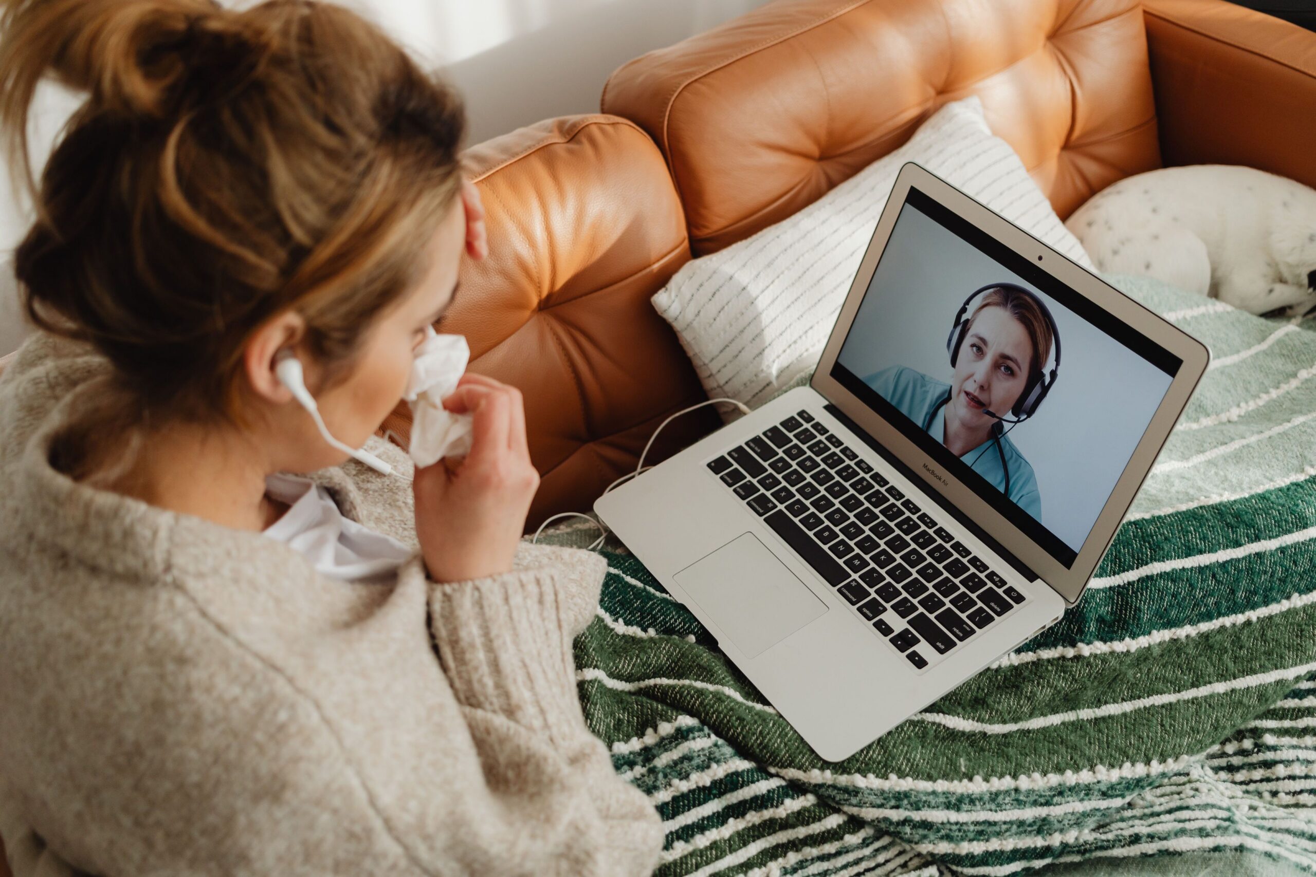 A woman with a cold sits on her sofa while on a telemed video call with her healthcare professional.