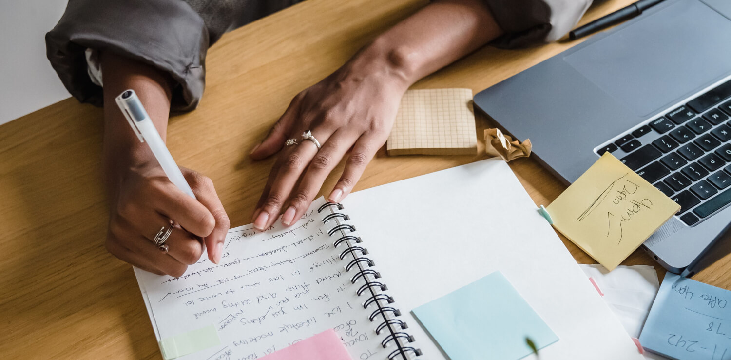 Therapist writing client notes at a desk covered in messy notes next to a laptop.
