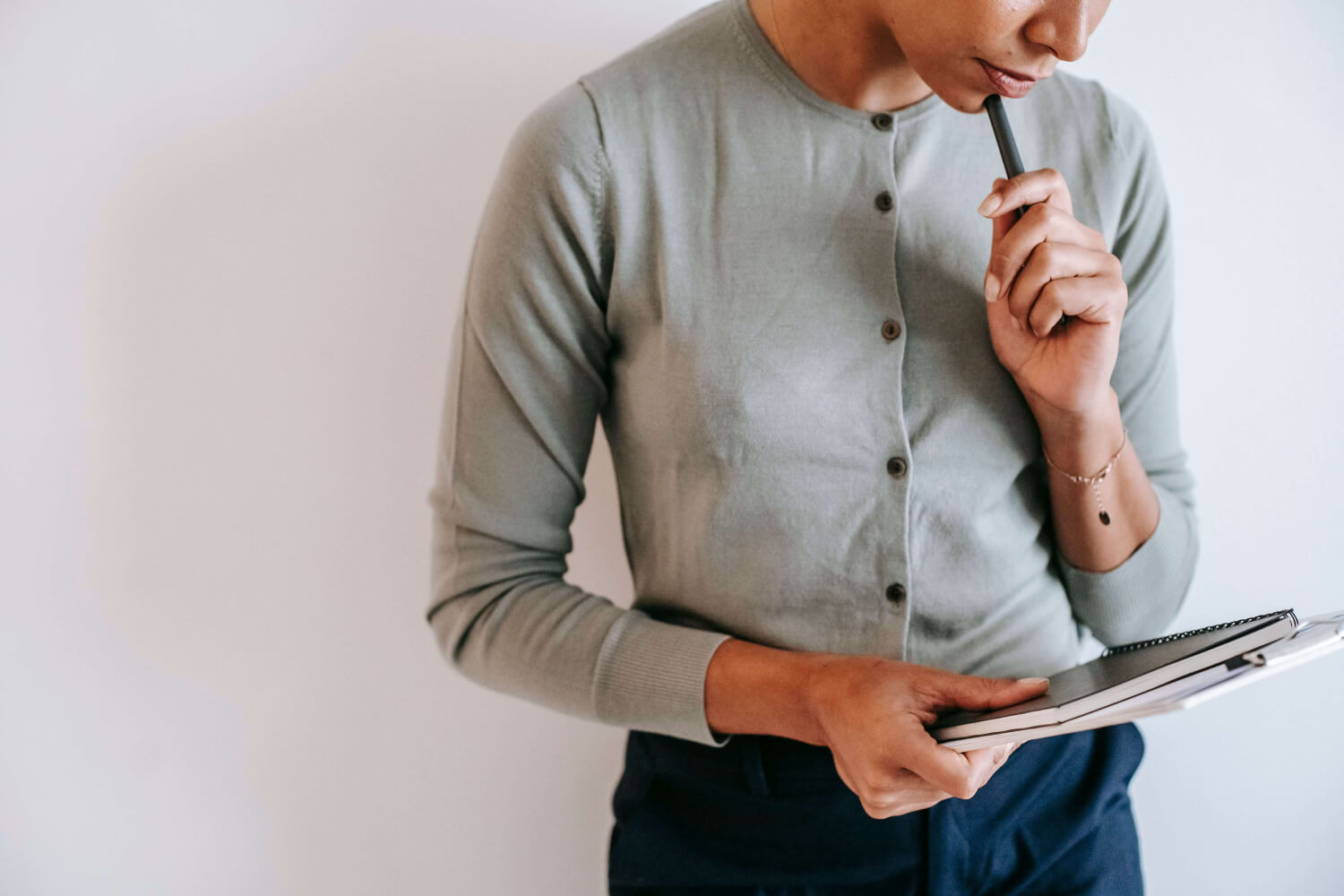 Therapist in a cardigan standing in front of a white wall looking down thoughtfully at the clipboard, notes and notebook in her hand.