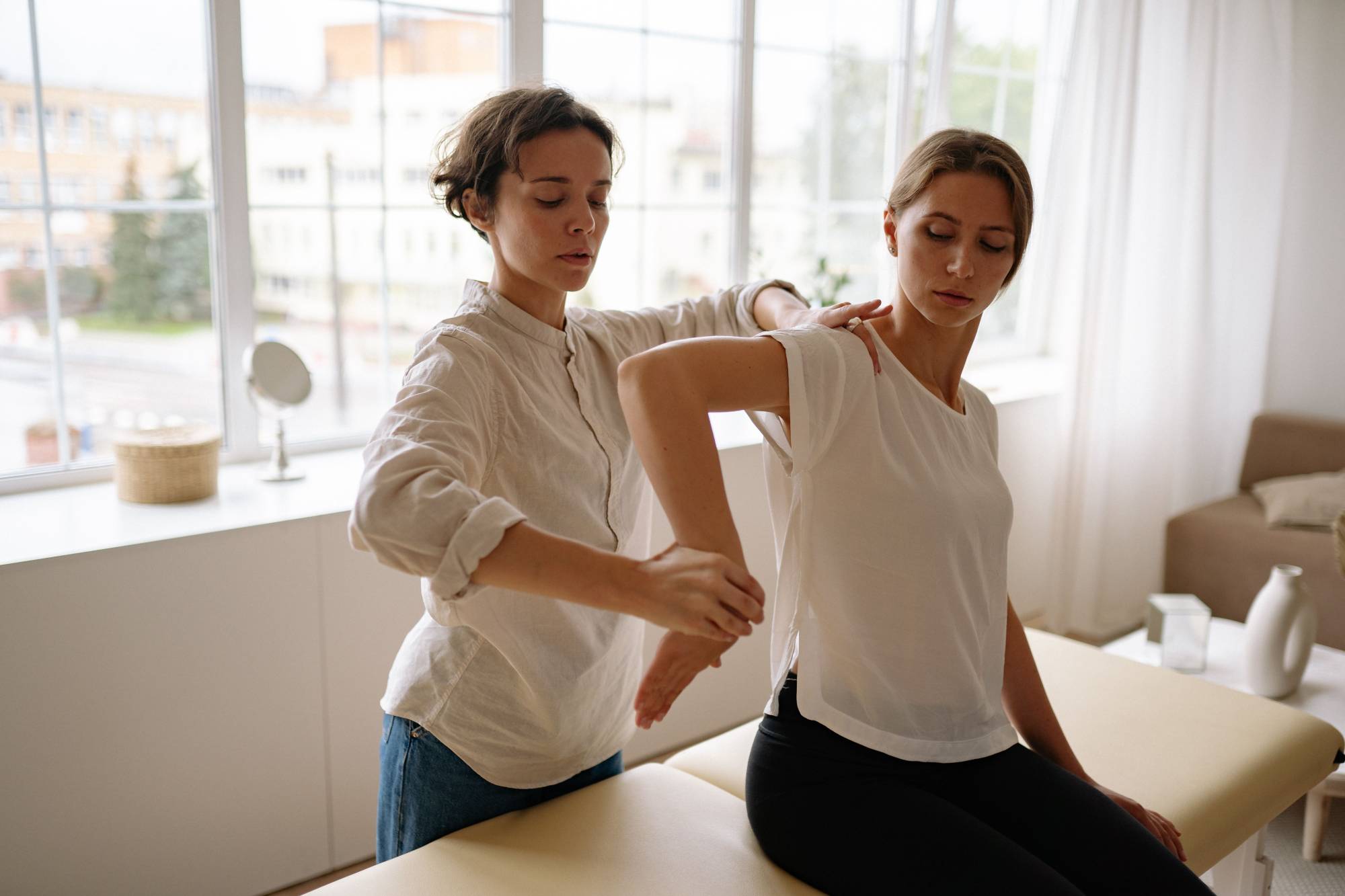 A healthcare practitioner treats a client’s shoulder while on an exam table in a clinic with big windows behind them.