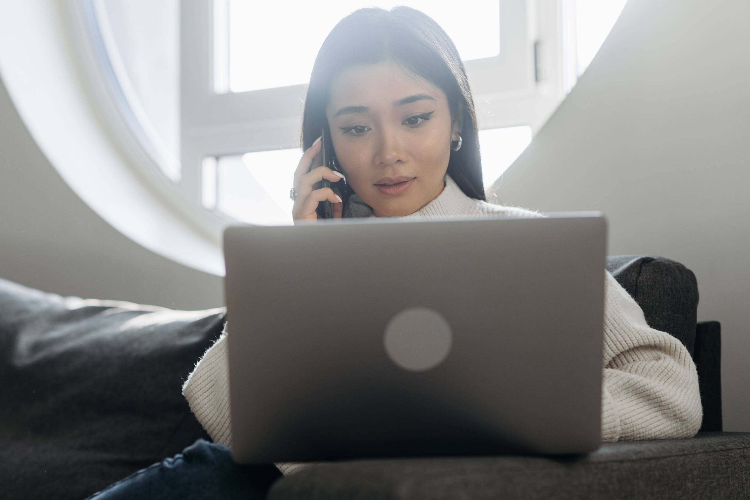 Image of a woman sits in front of a bright window as she talks on the phone and looks at her laptop.